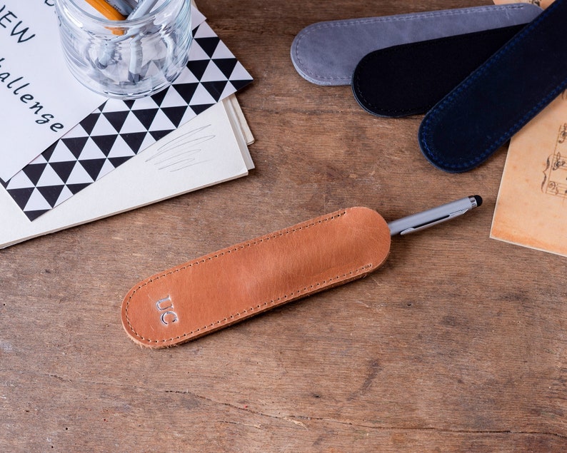 May include: A brown leather pen case with a silver pen inside. The case has the initials "UC" embossed on the front.