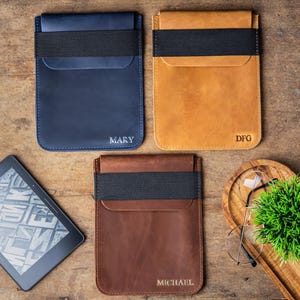 May include: Three leather e-reader sleeves in navy blue, tan, and brown, each with a black elastic band and personalized text. An e-reader is visible on the left. A wooden tray with glasses and a plant is on the right. The sleeves are on a wooden surface.