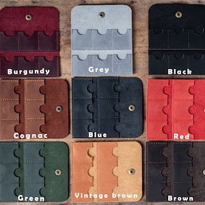 May include: Nine leather card holders in different colours, including burgundy, grey, black, cognac, blue, red, green, vintage brown, and brown. Each holder has six slots for cards.