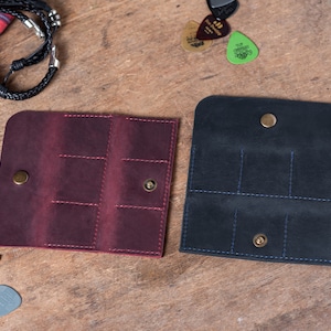 May include: Two leather guitar pick holders, one burgundy and one black, with multiple slots for picks and a snap closure.
