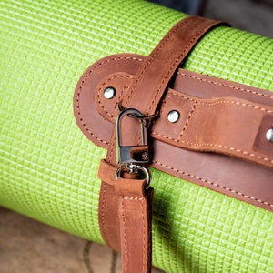 May include: A green yoga mat with a brown leather strap and a silver metal hook. The strap has stitching and rivets.