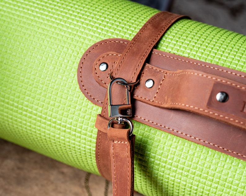 May include: A green yoga mat with a brown leather strap and a silver metal hook. The strap has stitching and rivets.