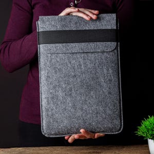 May include: A rectangular grey felt laptop sleeve featuring a horizontal black elastic band across the top flap. The sleeve has a structured design with stitched edges and a smooth, uniform texture, providing a protective cover for a portable computer.