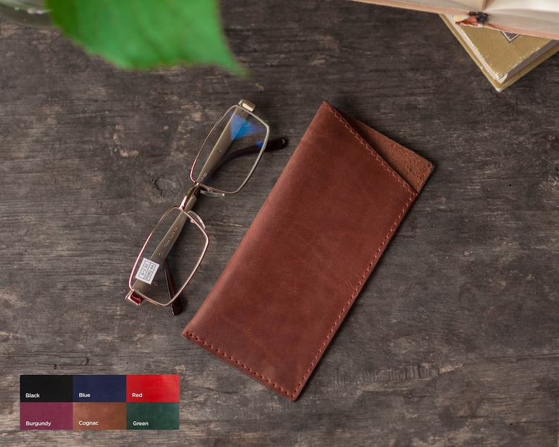 May include: A brown leather glasses case with a flap closure. The case is lying on a wooden surface next to a pair of clear glasses.
