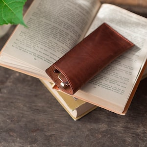 May include: A brown leather glasses case resting on top of a stack of books. The case is closed and has a subtle sheen.