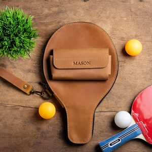 May include: A brown leather ping pong paddle case with a matching wallet, both with the name "MASON" embossed. Two yellow ping pong balls, a white ping pong ball, and a red and black ping pong paddle are also visible on a wooden surface.