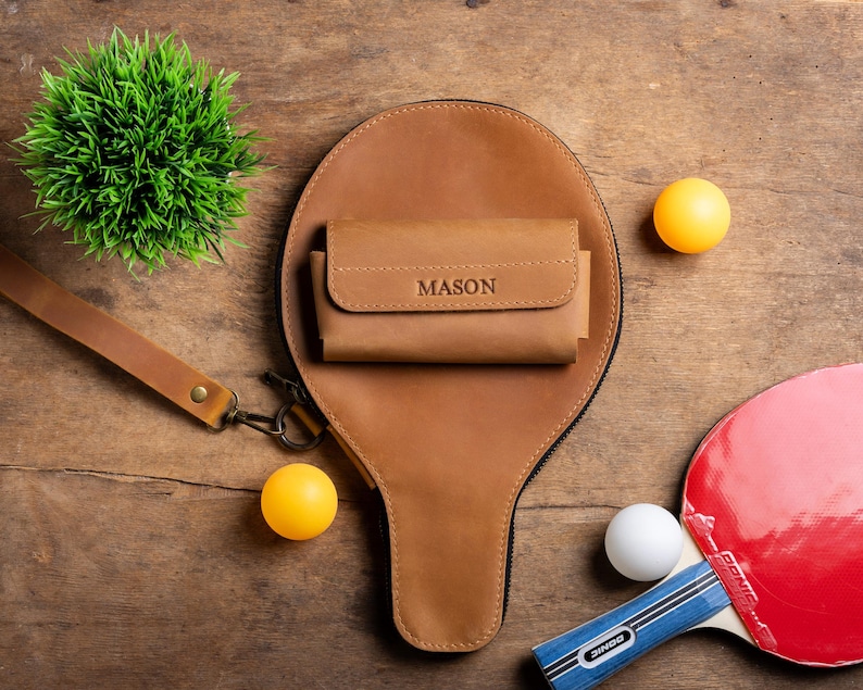 May include: A brown leather ping pong paddle case with a matching wallet, both with the name "MASON" embossed. Two yellow ping pong balls, a white ping pong ball, and a red and black ping pong paddle are also visible on a wooden surface.