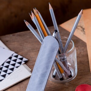 May include: A grey suede pencil case with a zip closure. The case is lying on a wooden surface next to a glass jar filled with pencils and pens.