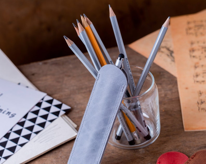 May include: A grey suede pencil case with a zip closure. The case is lying on a wooden surface next to a glass jar filled with pencils and pens.