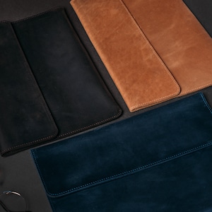 May include: Three leather laptop sleeves in different colors: black, brown, and blue. The sleeves are laid out on a dark surface.