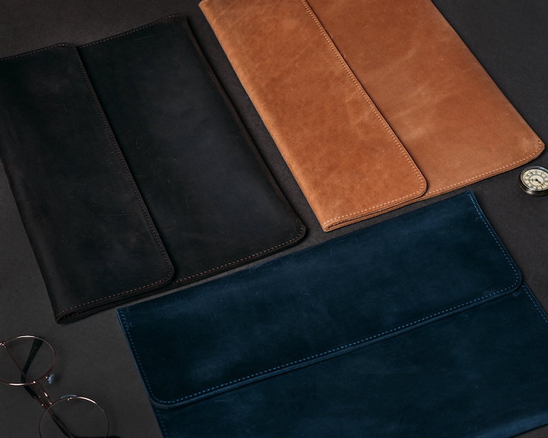 May include: Three leather laptop sleeves in different colors: black, brown, and blue. The sleeves are laid out on a dark surface.
