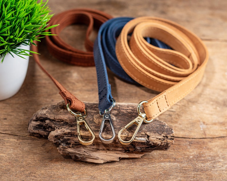 May include: Three leather dog leads in brown, tan, and blue with gold metal clips. The leads are coiled and arranged on a piece of driftwood.