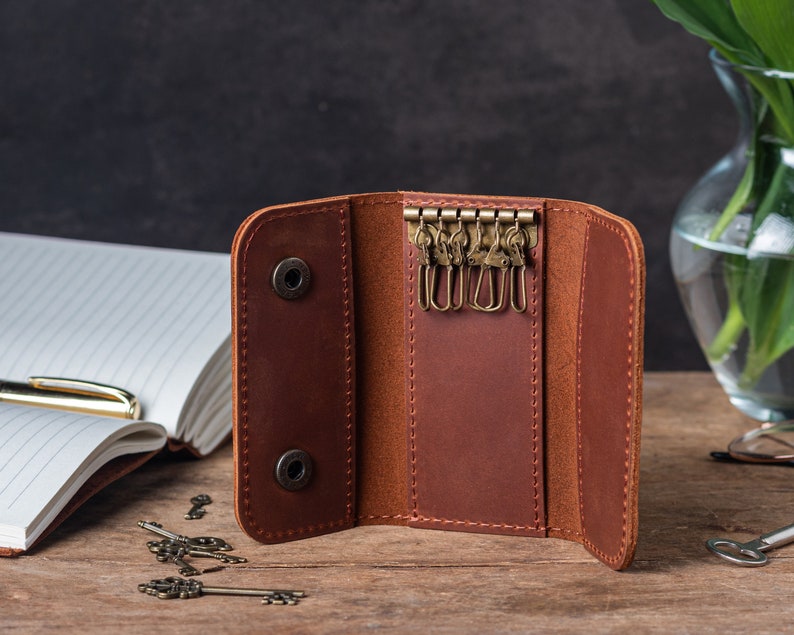 May include: A brown leather key holder with six key rings and two snap closures. The key holder is open and lying on a wooden surface.