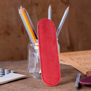 May include: A red leather pencil case with a stitched edge. The case is sitting on a wooden table next to a glass jar filled with pencils.