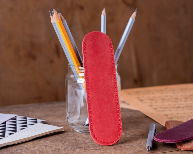 May include: A red leather pencil case with a stitched edge. The case is sitting on a wooden table next to a glass jar filled with pencils.