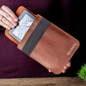 May include: A brown leather e-reader case with the name "MICHAEL" embossed on the front. The case has a black elastic band and an orange interior. The e-reader is partially visible inside the case.