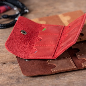 May include: A red leather guitar pick holder with multiple slots for storing picks. The holder is open and folded back to reveal the interior slots. The holder is made of a soft, textured leather.