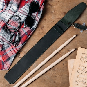 Puede incluir: Un porta baquetas de cuero verde oscuro con un llavero de metal y un par de baquetas de madera. El porta baquetas est&aacute; sobre una superficie de madera con una camisa de cuadros rojos y blancos y partituras musicales.