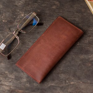 May include: A brown leather glasses case with a stitched edge. The case is lying on a wooden surface next to a pair of silver-rimmed glasses.