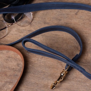 May include: A blue leather dog leash with a gold metal clasp. The leash is lying on a wooden surface.