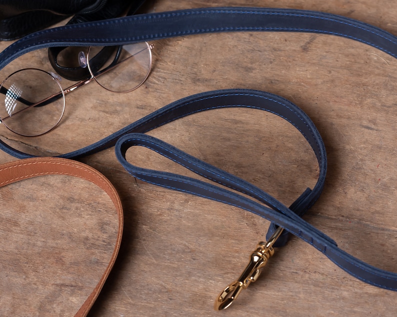May include: A blue leather dog leash with a gold metal clasp. The leash is lying on a wooden surface.