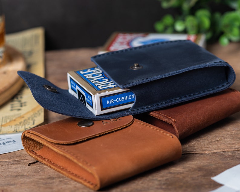 May include: Two leather card cases, one blue and one brown, with a deck of Bicycle playing cards inside the blue case. The blue case has a flap that closes with a snap. The brown case has a flap that closes with a snap and a small metal button. The deck of cards has the words "Bicycle Air-Cushion" printed on the side.