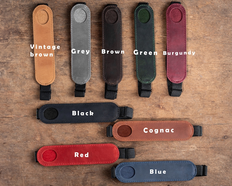 K&ouml;nnte beinhalten: Neun Leder-Kameragurtenden in verschiedenen Farben: Vintage Braun, Grau, Braun, Gr&uuml;n, Burgunder, Schwarz, Cognac, Rot und Blau. Jede Gurtenden hat ein rundes Loch in der Mitte.
