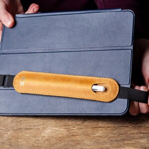 May include: A blue tablet case with a tan leather hand strap. The strap has the initials "H R" and a black elastic band. A white stylus is inserted into the leather strap. The case is held by two hands.