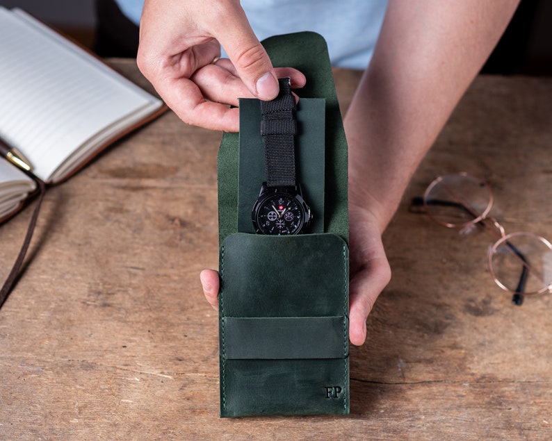 May include: A dark green leather watch case with a black watch inside. The case has a flap that folds over the watch and a small pocket on the front. The case has the initials "FP" embossed on the front.