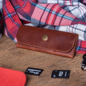 May include: A brown leather key holder with a gold button closure. The key holder is lying on a wooden surface next to a red and black checked fabric.
