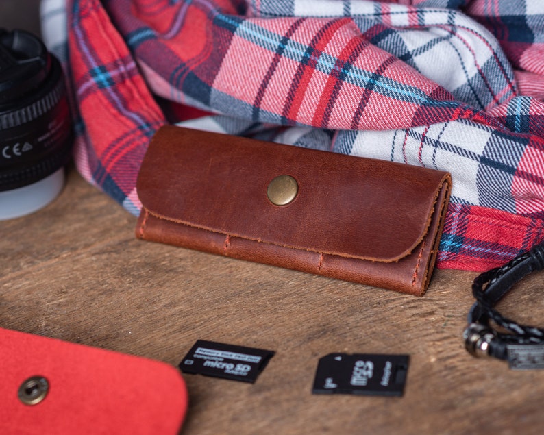 May include: A brown leather key holder with a gold button closure. The key holder is lying on a wooden surface next to a red and black checked fabric.
