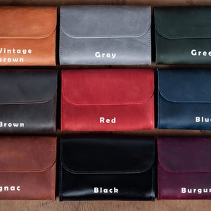 Op de afbeelding: Negen leren portemonnees in verschillende kleuren, waaronder vintage bruin, grijs, groen, bruin, rood, blauw, cognac, zwart en bordeauxrood. Elke portemonnee heeft een klep met een gestikte rand en een wit tekstlabel met de kleurnaam.
