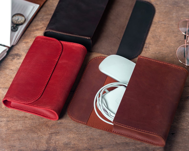 May include: A collection of leather cases in various colors, including red and brown. One case is open, revealing a white computer mouse and charging cable. The cases are arranged on a wooden surface, suggesting a theme of organization and storage.