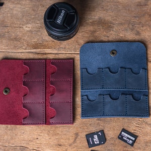 May include: Two leather cases for memory cards. The case on the left is burgundy and the case on the right is blue. Both cases have multiple slots for memory cards.