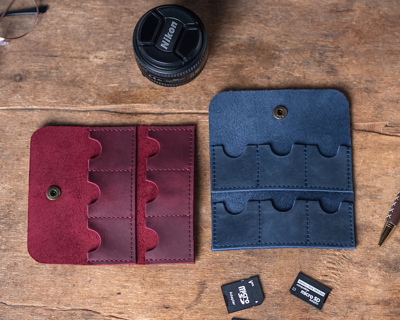 May include: Two leather cases for memory cards. The case on the left is burgundy and the case on the right is blue. Both cases have multiple slots for memory cards.