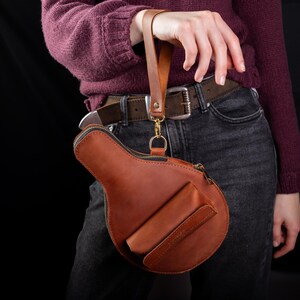 May include: A brown leather handbag with a unique shape, featuring a zipper closure and a small front pocket. The bag has a wrist strap and a gold-colored clasp. The bag is held by a person wearing a burgundy sweater and dark jeans.