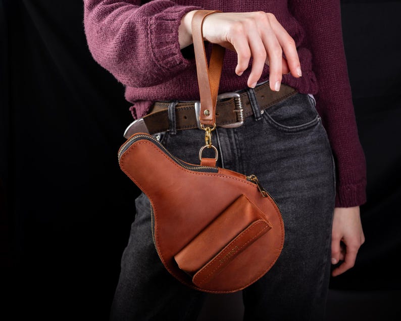 May include: A brown leather handbag with a unique shape, featuring a zipper closure and a small front pocket. The bag has a wrist strap and a gold-colored clasp. The bag is held by a person wearing a burgundy sweater and dark jeans.
