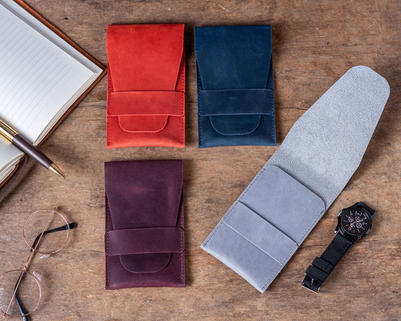 May include: Four leather watch cases in red, blue, gray, and burgundy. The cases are made of soft leather and have a flap that closes with a snap. The cases are all laid out on a wooden surface.