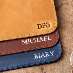May include: Close-up of three personalized leather cases in tan, brown, and blue. The tan case has the initials "DFG", the brown case says "MICHAEL", and the blue case says "MARY". Each case has a stitched border.