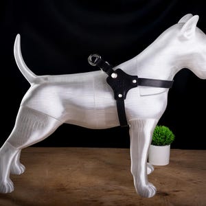 May include: A white dog statue with a black leather harness. The dog is standing on a wooden surface with a small green plant in a white pot in the background. The harness has a metal ring.