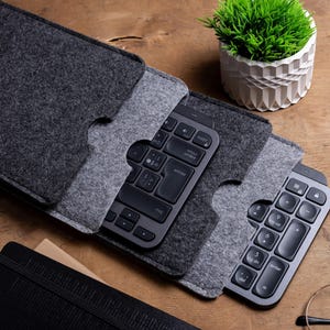 Puede incluir: Primer plano de dos teclados inalámbricos en fundas de fieltro gris. Los teclados son negros con teclas grises. Una pequeña maceta y un cuaderno negro también son visibles en la superficie de madera.