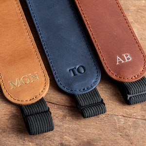 K&ouml;nnte beinhalten: Drei Lederkameragurte mit personalisierten Initialen. Die Gurte sind braun, blau und beige. Jeder Gurt hat ein schwarzes Gummiband.