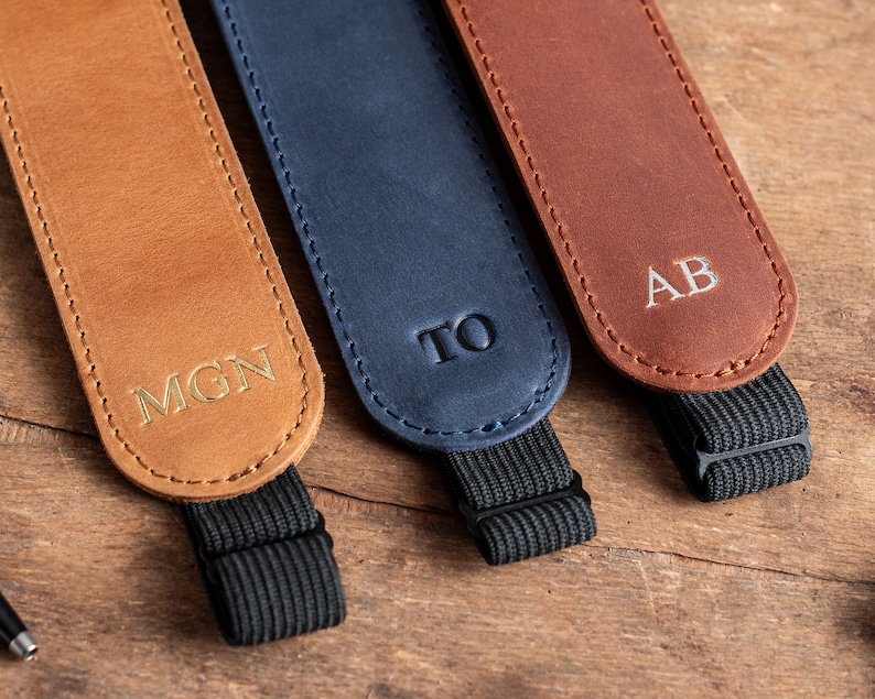 K&ouml;nnte beinhalten: Drei Lederkameragurte mit personalisierten Initialen. Die Gurte sind braun, blau und beige. Jeder Gurt hat ein schwarzes Gummiband.