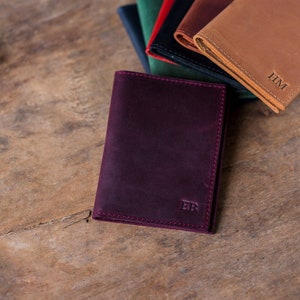 May include: A burgundy leather passport holder with the initials "EB" embossed on the front. The holder is lying on a wooden surface with other leather passport holders in the background.
