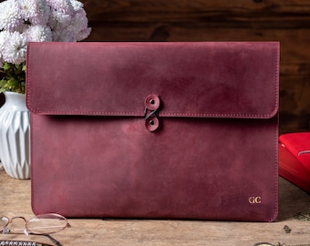 May include: A burgundy leather laptop sleeve with a flap closure and two black button accents. The sleeve has a gold embossed "GC" monogram on the bottom right corner.