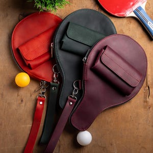 May include: A collection of leather ping pong paddle-shaped bags in red, dark green, and burgundy, each with a small pocket and a wrist strap. A red and blue ping pong paddle and two ping pong balls are also visible.