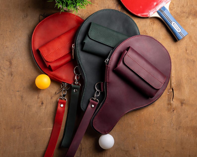 May include: A collection of leather ping pong paddle-shaped bags in red, dark green, and burgundy, each with a small pocket and a wrist strap. A red and blue ping pong paddle and two ping pong balls are also visible.