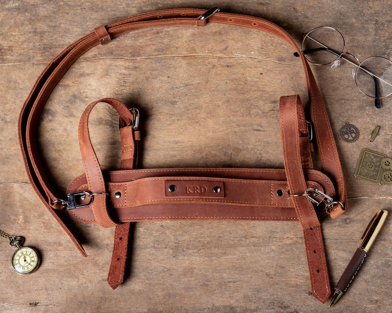 May include: A brown leather camera strap with adjustable straps and a buckle. The strap has the word "KRD" embossed on it.
