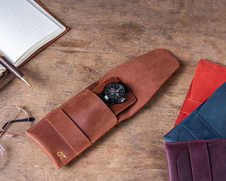 May include: A brown leather watch case with a watch inside. The case has the initials "GT" embossed on the front. There are other leather cases in red, blue, and purple in the background.