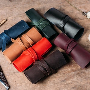 May include: Seven leather rolls in various colors, including brown, red, black, green, and blue. Each roll has a leather strap with stitching and is rolled up with a label that says "BY".